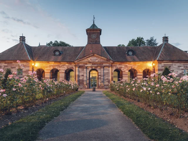 Vue extérieure du bâtiment des thermes de Luxeuil-les-Bains à la tombée de la nuit, construit en grès rose des Vosges et dont l'allée est bordée de rosiers - Vosges du Sud