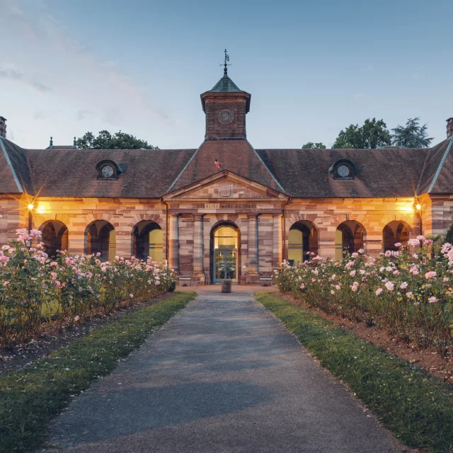 Vue extérieure du bâtiment des thermes de Luxeuil-les-Bains à la tombée de la nuit, construit en grès rose des Vosges et dont l'allée est bordée de rosiers - Vosges du Sud