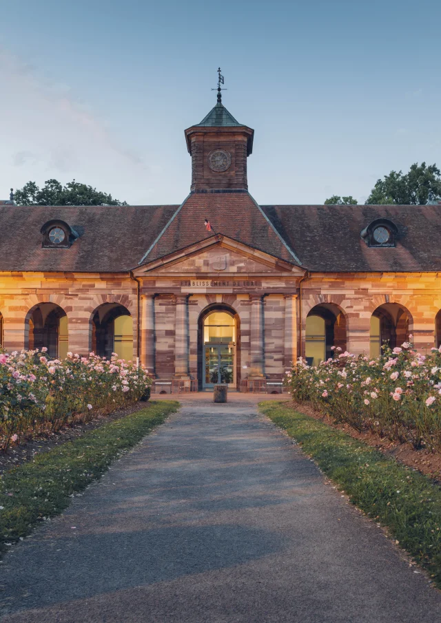 Vue extérieure du bâtiment des thermes de Luxeuil-les-Bains à la tombée de la nuit, construit en grès rose des Vosges et dont l'allée est bordée de rosiers - Vosges du Sud