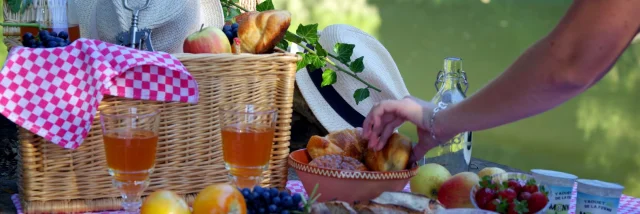 Une femme se sert dans un panier de pique nique près de l'eau. Sur une nappe à carreaux roses se trouvent des produits locaux : terrine, fromage, fruits, laitage, viennoiserie ...