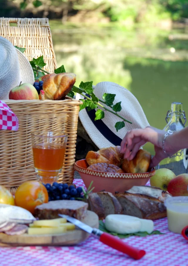 Une femme se sert dans un panier de pique nique près de l'eau. Sur une nappe à carreaux roses se trouvent des produits locaux : terrine, fromage, fruits, laitage, viennoiserie ...