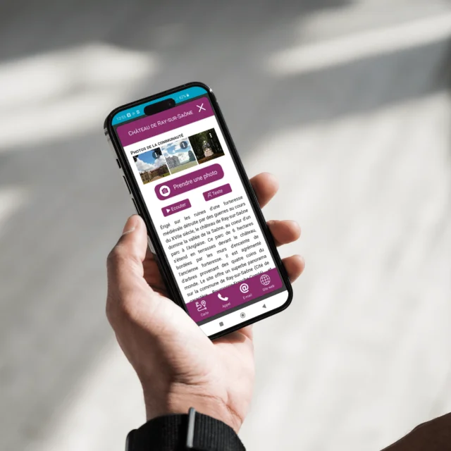 Une personne tient son smartphone avec l'application My Haute-Saône ouverte sur la page du Château de Ray-sur-Saône avec la possibilité de partager ses propres photos