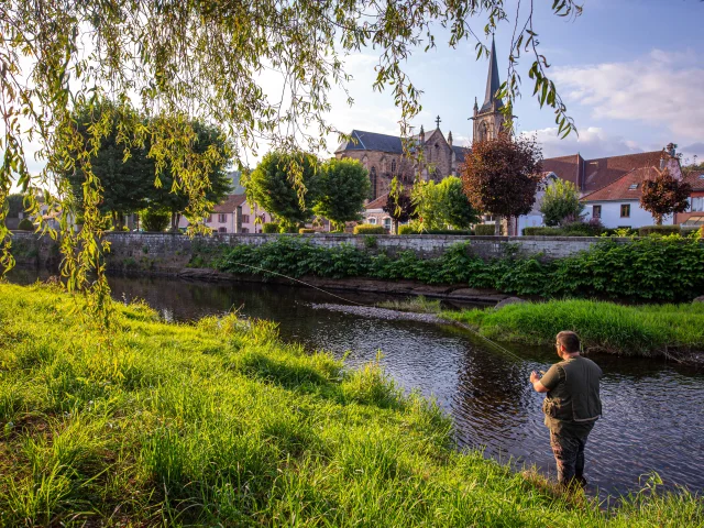 Une femme et un homme pêche dans le Rahin à Ronchamp. À l'arrière-plan, on observe la commune avec son église Notre-Dame du Bas - Vosges du Sud