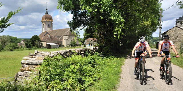 Un groupe de VTTistes arpente les Monts de Gy avec une église aux tuiles vernissées en arrière-plan - Vallée de l'Ognon