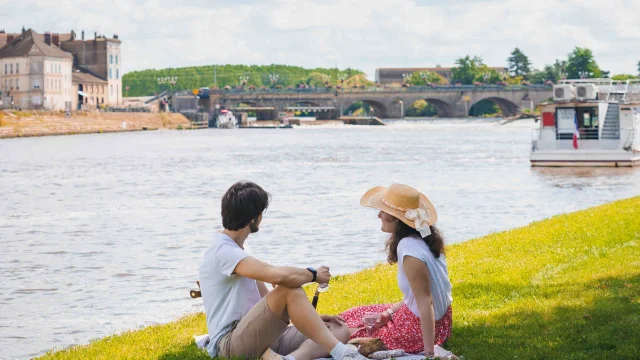 Un couple pique-nique, dans l'herbe, sur les bords de Saône à Gray. A l'arrière-plan on voit un bateau de plaisance, un pont et des bâtiments - Gray - Vesoul - Val de Saône