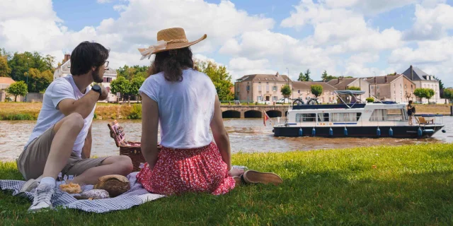 Un couple pique-nique, dans l'herbe, sur les bords de Saône à Gray. A l'arrière-plan on voit un bateau de plaisance en train de navigeur, un pont et des bâtiments - Gray - Vesoul - Val de Saône