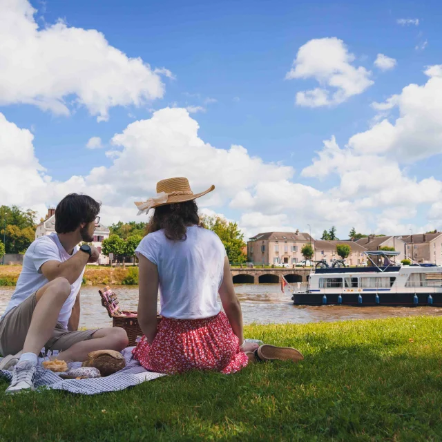 Un couple pique-nique, dans l'herbe, sur les bords de Saône à Gray. A l'arrière-plan on voit un bateau de plaisance en train de navigeur, un pont et des bâtiments - Gray - Vesoul - Val de Saône