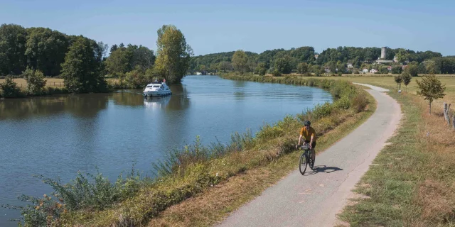 Un cyclise itinérant se déplace sur la Voie Bleue, tandis qu'un bateau navigue sur la Saône. Au fond, on retrouve le village de Rupt-sur-Saône et son château - Vesoul - Val de Saône