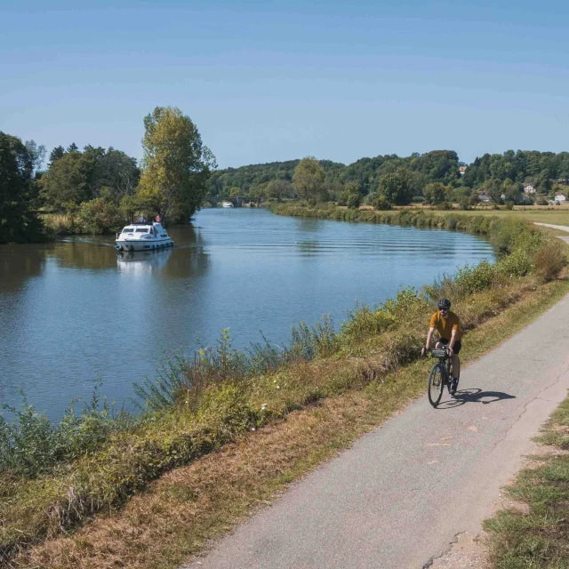 Un cyclise itinérant se déplace sur la Voie Bleue, tandis qu'un bateau navigue sur la Saône. Au fond, on retrouve le village de Rupt-sur-Saône et son château - Vesoul - Val de Saône