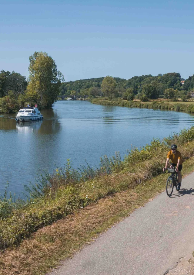 Un cyclise itinérant se déplace sur la Voie Bleue, tandis qu'un bateau navigue sur la Saône. Au fond, on retrouve le village de Rupt-sur-Saône et son château - Vesoul - Val de Saône