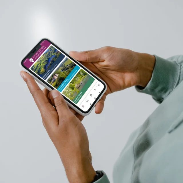 Une personne tient un smartphone avec l'application My Haute-Saône ouverte sur la page d’accueil, présentant les 3 destinations : Vesoul-Val de Saône, Vosges du Sud et Vallée de l’Ognon - Haute-Saône