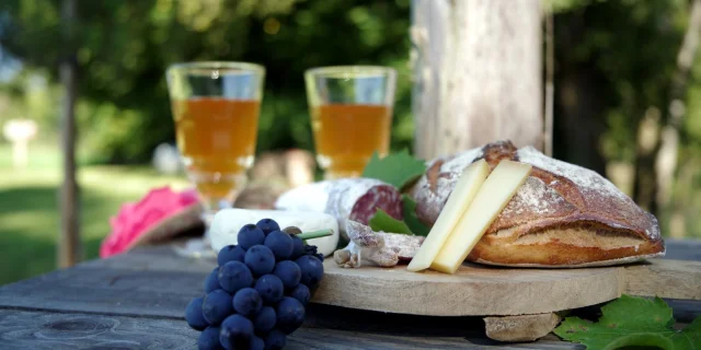 Plateau avec du pain, du fromage, saucisson, 2 verres de jus de pommes. L'image est colorée avec un chapeau rose, du raisin et du lierre.