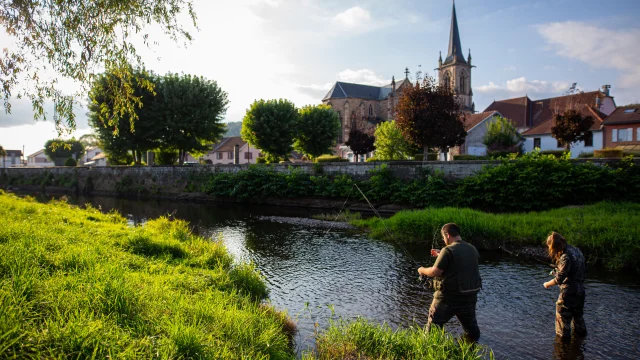 Une femme et un homme sont en train de pêcher aux leurres dans le Rahin à Ronchamp. À l'arrière-plan, on observe la commune avec son église Notre-Dame du Bas - Vosges du Sud