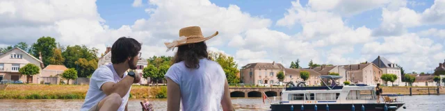 Un couple pique-nique, dans l'herbe, sur les bords de Saône à Gray. A l'arrière-plan on voit un bateau de plaisance en train de navigeur, un pont et des bâtiments - Gray - Vesoul - Val de Saône