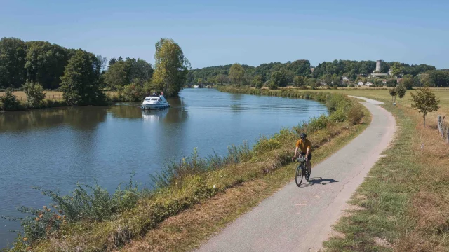 Un cyclise itinérant se déplace sur la Voie Bleue, tandis qu'un bateau navigue sur la Saône. Au fond, on retrouve le village de Rupt-sur-Saône et son château - Vesoul - Val de Saône