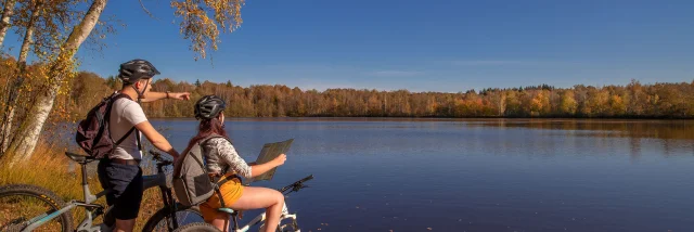 Un couple en VTT fiat une pause au bord d'un étang, avec des couleurs automnales, sur le Plateau des 1000 Etangs - Vosges du Sud