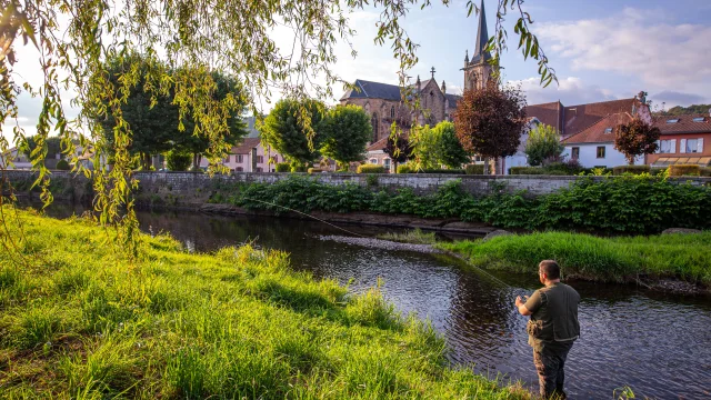 Une femme et un homme pêche dans le Rahin à Ronchamp. À l'arrière-plan, on observe la commune avec son église Notre-Dame du Bas - Vosges du Sud