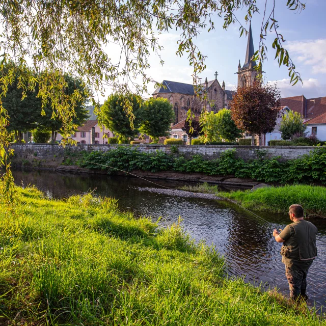 Une femme et un homme pêche dans le Rahin à Ronchamp. À l'arrière-plan, on observe la commune avec son église Notre-Dame du Bas - Vosges du Sud